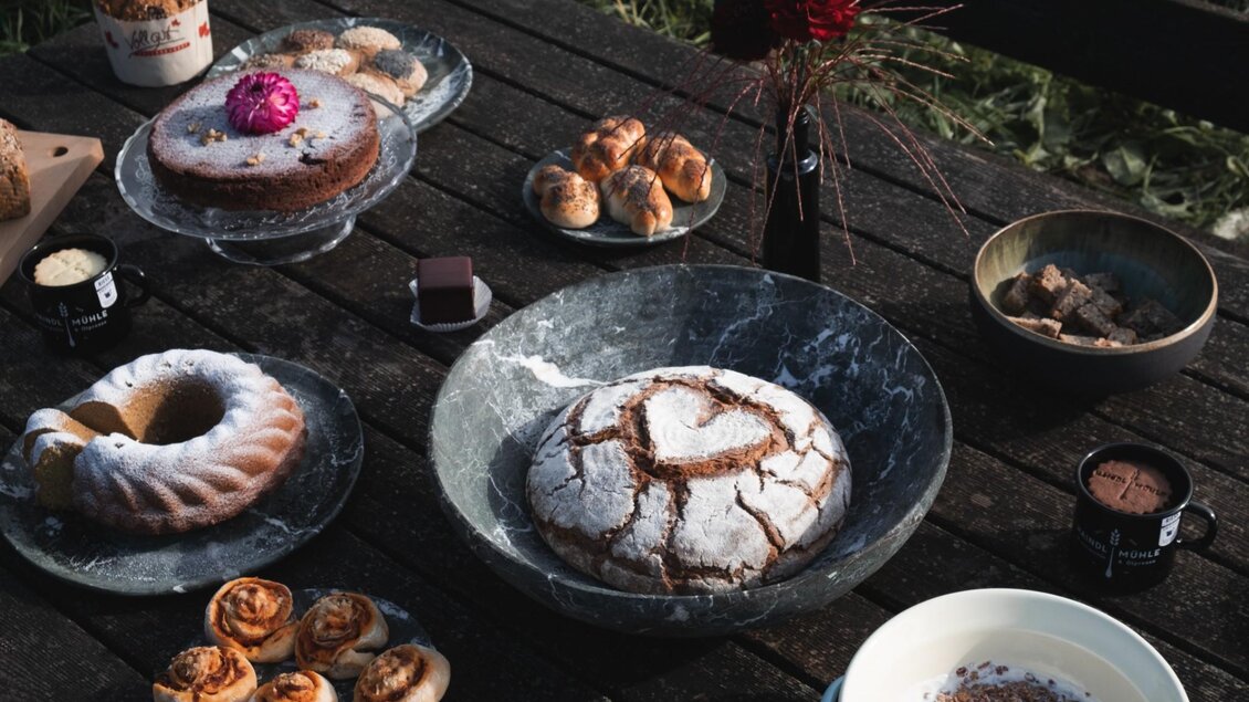 Vielfältige Brote, Kuchen und Gebäck auf Holztisch vor Wiese, Haindl-Mühle-Tasse sichtbar | © ee photograph - Elena Egger