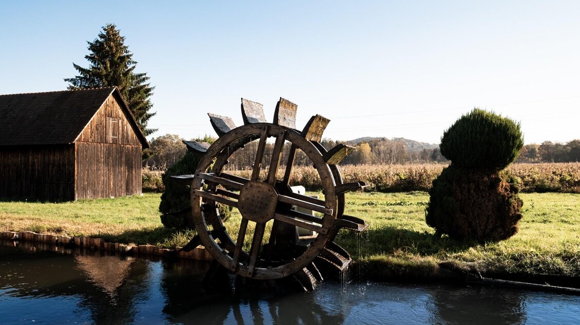 Großes hölzernes Wasserrad am Wasserlauf vor Wiese und Holzhütte bei der Haindl-Mühle | © ee photograph - Elena Egger