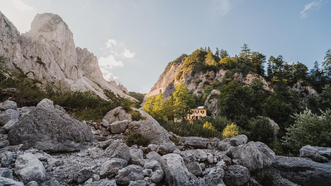 Eine malerische Berglandschaft mit hohen Felsen und grünen Bäumen. Im Hintergrund ist ein kleines Haus zu sehen, umgeben von Natur.