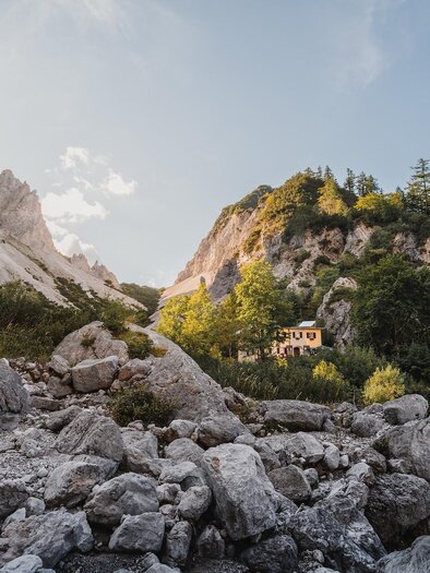 A picturesque mountain landscape with high cliffs and green trees. In the background, a small house can be seen, surrounded by nature.