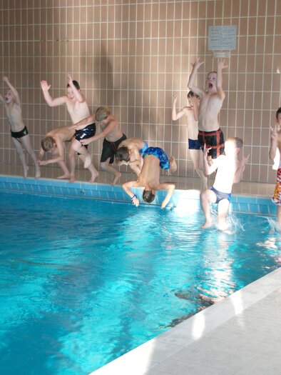 A group of children joyfully jumps into a swimming pool. The water is clear and inviting, and the sun shines through the windows. | © Gemeinde Pöls-Oberkurzheim