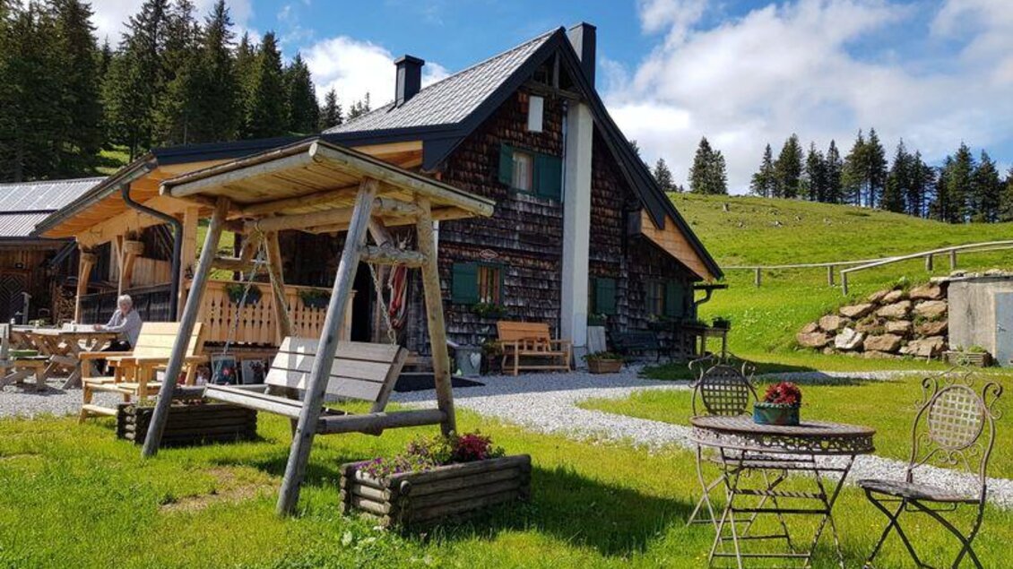 Eine gemütliche Berghütte umgeben von grünen Wiesen und Bäumen. Im Vordergrund stehen eine Schaukel und ein Tisch mit Stühlen. | © Rotsohlalm