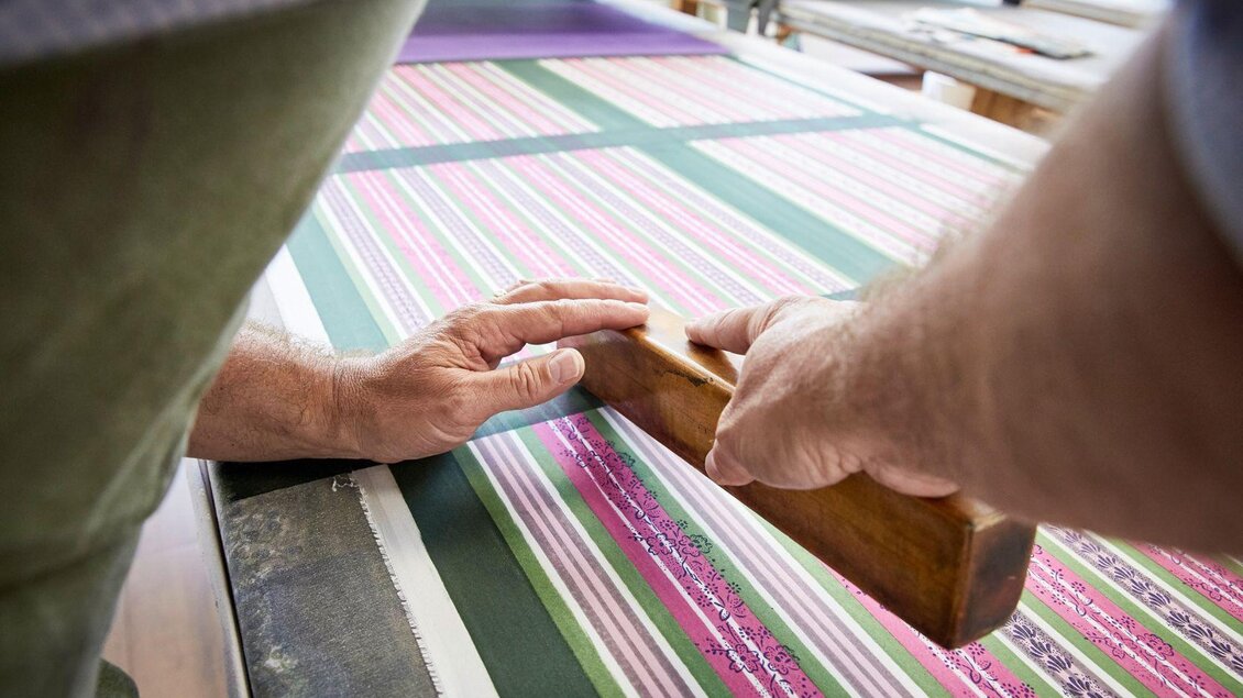 Hände arbeiten an einem Drucktisch mit buntem Streifenmuster. Der Fokus liegt auf der Drucktechnik mit einem Holzdruckwerkzeug. | © Handdrucke Sekyra-Helge Kirchberger
