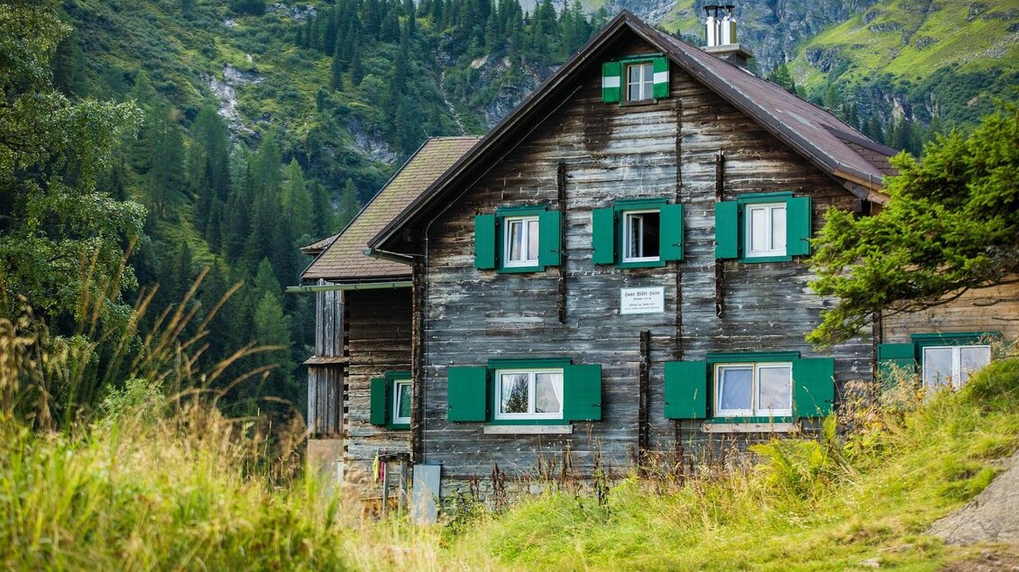Ein rustikales Holzhaus in der Natur, umgeben von grünen Wäldern und Bergen. Die Fensterläden sind grün gestrichen und verleihen dem Gebäude einen charmanten Look. | © TVB Haus-Aich-Gössenberg@René Eduard Perhab
