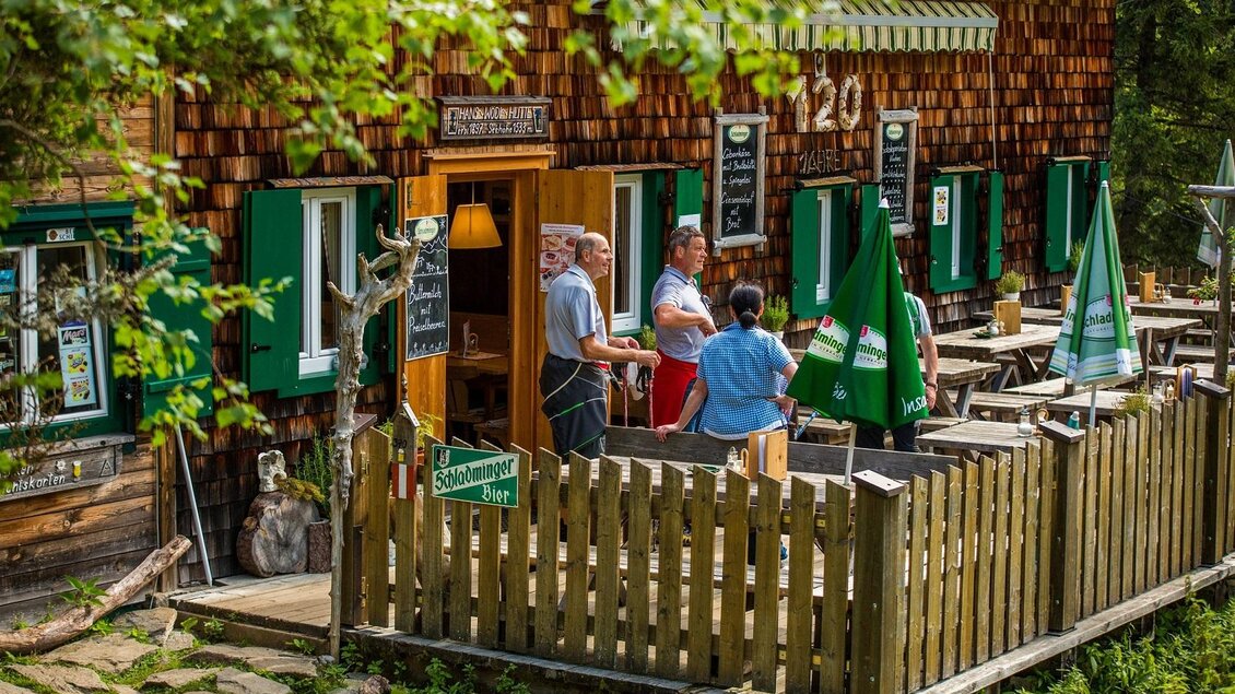 Ein gemütliches Restaurant im Freien mit Holzverkleidung und Tischen auf einer Terrasse. Gäste stehen im Gespräch vor dem Eingang, umgeben von grünen Bäumen und Pflanzen. | © TVB Haus-Aich-Gössenberg@René Eduard Perhab