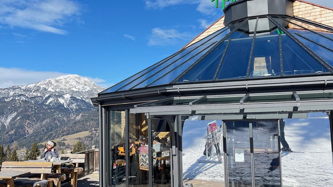 Ein modernes Alpengasthaus mit Glasdach liegt in einer schneebedeckten Landschaft. Im Hintergrund sind majestätische Berge und ein blauer Himmel zu sehen. | © Keinprecht