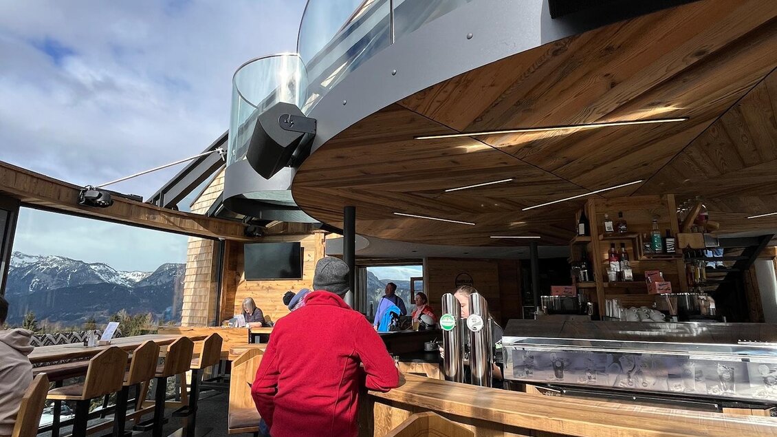 Eine moderne Bar mit einem Holzinterieur und einer großen Glasfront, die einen schönen Ausblick auf die Berge bietet. Im Vordergrund ist eine Person in einem roten Oberteil beim Bestellen. | © Keinprecht