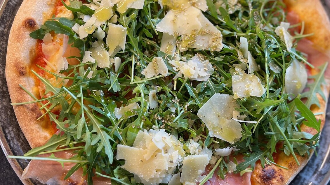 Eine Pizza mit frischem Rucola, Schinken und Parmesanstücken. Der Teig ist knusprig und goldbraun. | © Keinprecht