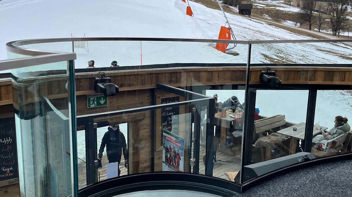 Eine moderne Aussichtsterrasse mit Glaswänden bietet einen Blick auf eine verschneite Skipiste. Im Hintergrund sind Skifahrer und eine winterliche Landschaft zu sehen. | © Keinprecht