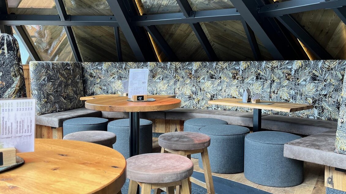 Ein modernes Café mit großen Fenstern und gemütlichen Sitzmöglichkeiten. Holz- und Stoffelemente schaffen eine einladende Atmosphäre. | © Keinprecht