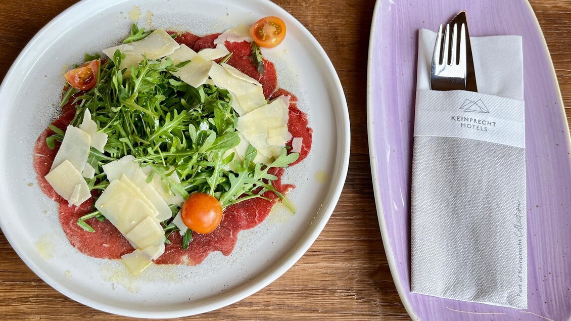 Ein Teller mit Carpaccio, garniert mit Rucola, Parmesan und Kirschtomaten. Daneben liegt ein Besteckset auf einem eleganten Teller. | © Keinprecht
