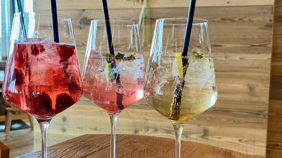 Drei farbenfrohe Cocktails in eleganten Gläsern stehen auf einem Holztisch. Im Hintergrund ist ein Schild mit der Aufschrift "HAPPY ALM" sichtbar. | © Keinprecht