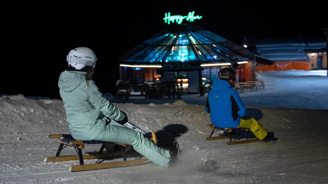 Zwei Personen rodeln im Schnee bei Nacht. Im Hintergrund ist ein beleuchtetes Gebäude mit dem Schriftzug "Happy Hour" zu sehen. | © Harald Steiner