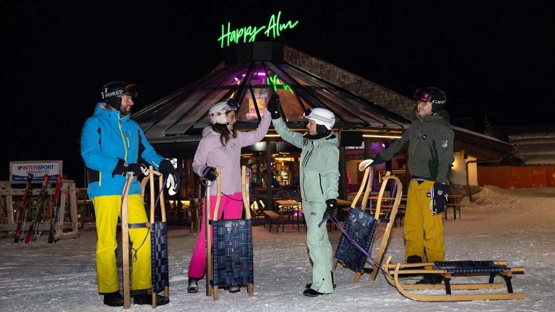 Eine Gruppe von Freunden steht draußen im Schnee vor einer Hütte mit dem Namen "Happy Alm". Sie tragen Winterkleidung und halten Schlitten in den Händen. | © Harald Steiner