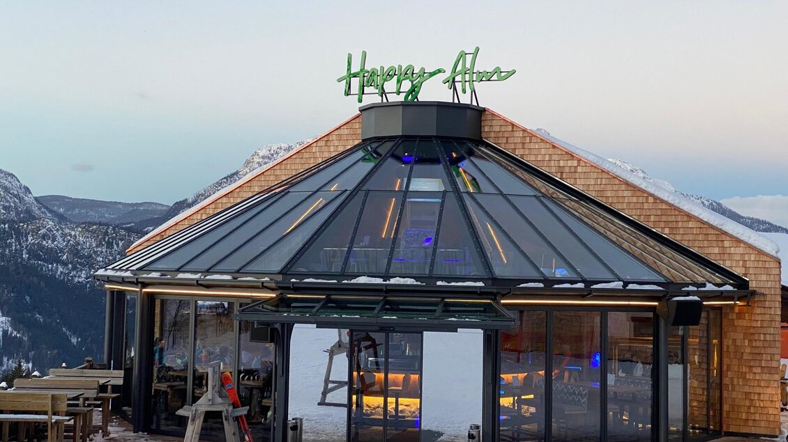 Ein modernes Restaurant mit einer gläsernen Fassade und einem einladenden Schild. Umgeben von schneebedeckten Bergen unter einem klaren Himmel. | © Happy Alm