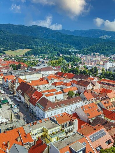 A picturesque view of a town with red roofs and green hills in the background. The architecture features a blend of historic and modern buildings. | © TV Erzberg Leoben