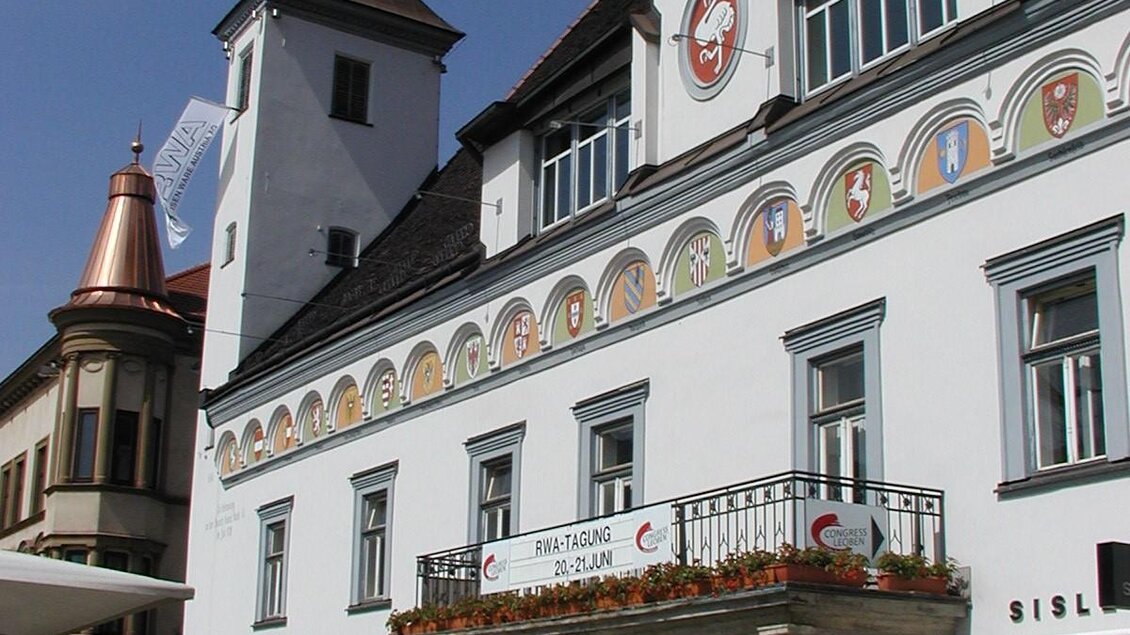 Ein historisches Gebäude mit einer Uhrturm und bunten Wandmalereien. Vor dem Gebäude sitzen Tische und Stühle im Freien. | © TV Erzberg Leoben