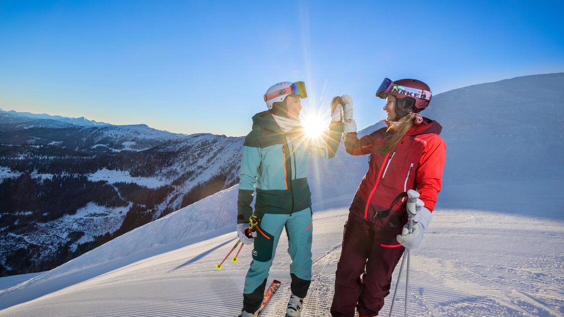 Zwei Skifahrer auf einer verschneiten Piste feiern den Sonnenuntergang. Im Hintergrund erstreckt sich eine wunderschöne Berglandschaft. | © Bernhard Moser