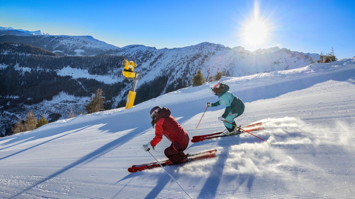 Zwei Skifahrer fahren die Piste hinunter, umgeben von schneebedeckten Bergen. Die Sonne scheint klar am Himmel und es gibt keine Wolken. | © Bernhard Moser