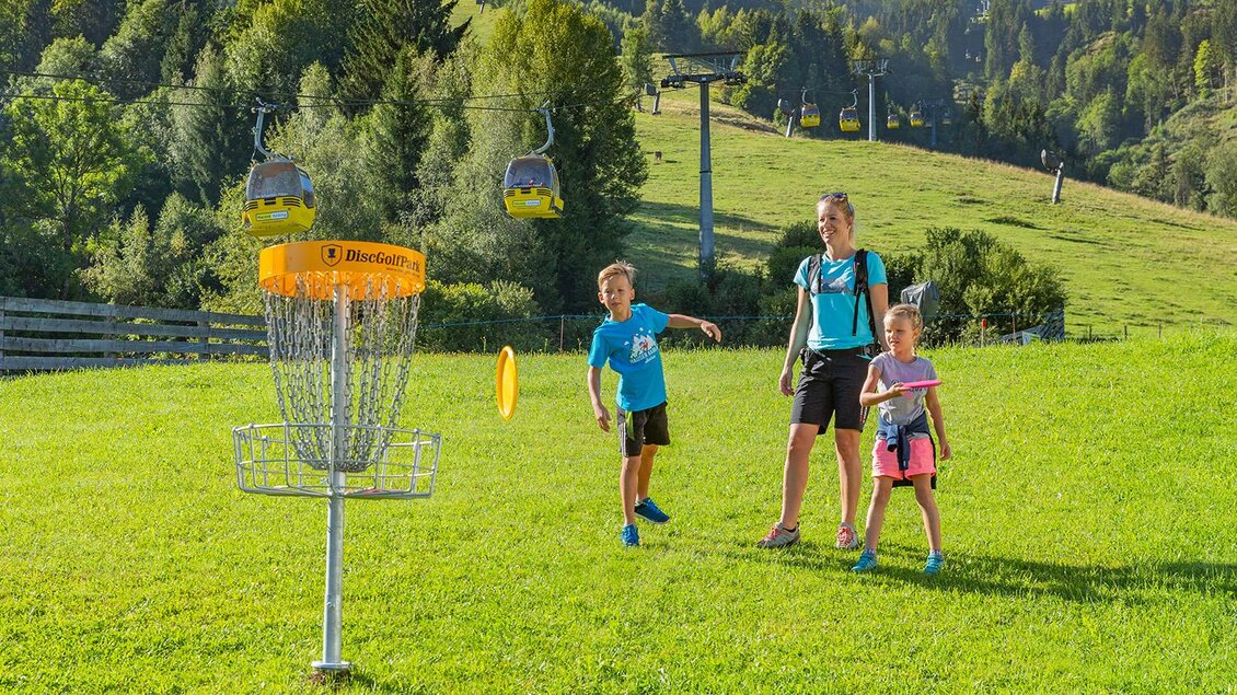 Ein Spielplatz mit einem Discgolf-Korb auf einer grünen Wiese. Zwei Kinder und eine Erwachsene stehen lachend, während eines der Kinder einen Wurf macht. | © Hauser Kaibling@René Eduard Perhab
