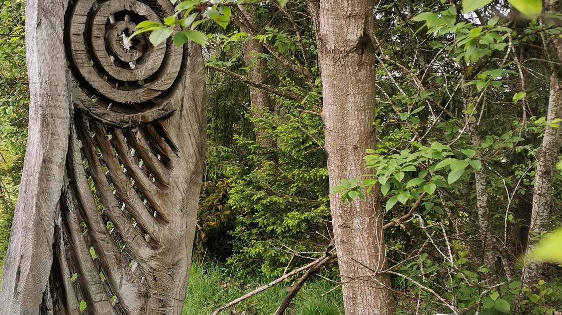 Eine Holzskulptur mit einem spiralförmigen Muster steht im Wald. Daneben befindet sich ein Informationsschild. | © Landentwicklung Steiermark