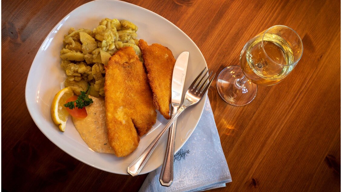 Ein gedeckter Teller mit knusprigem Fisch, Beilagen und einem Glas Wein. Dazu sieht man ein bisschen Zitronen- und Petersilien garnierung. | © TV Region Graz - Lunghammer