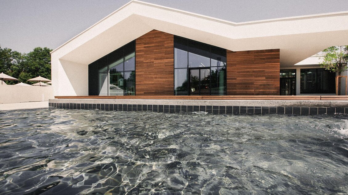 Heilthermen Resort Bad Waltersdorf: Ein modernes Gebäude mit großen Fenstern und Holzverkleidung steht an einem Pool. Das Wasser ist klar und reflektiert die Architektur. | © Heilthermen Resort Bad Waltersdorf