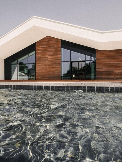 Healing Thermal Resort Bad Waltersdorf: A modern building with large windows and wood cladding stands by a pool. The water is clear and reflects the architecture. | © Heilthermen Resort Bad Waltersdorf