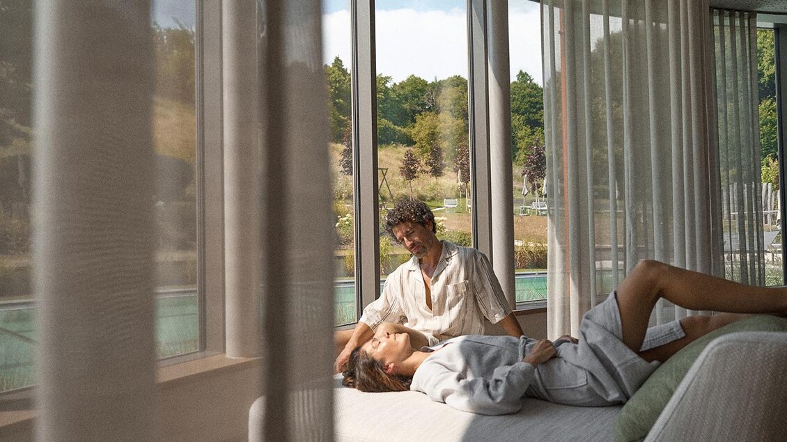 Ein Paar sitzt entspannt im Raum mit großen Fenstern. Licht strömt herein und schafft eine gemütliche Atmosphäre. | © Heilthermen Resort Bad Waltersdorf