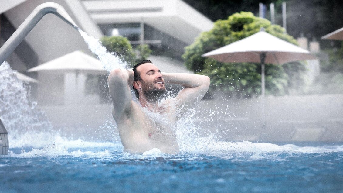 Ein Mann entspannt im Wasser eines Whirlpools. Über ihm sprühen Wasserdüsen und im Hintergrund sind Sonnenschirme und Grüne zu sehen. | © Heilthermen Resort Bad Waltersdorf