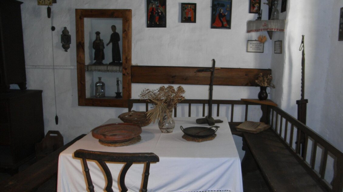 Ein traditionelles Zimmer mit einem Tisch und Bänken aus Holz. An den Wänden hängen Bilder und es gibt eine Vitrine mit dekorativen Objekten. | © Stadtgemeinde Feldbach