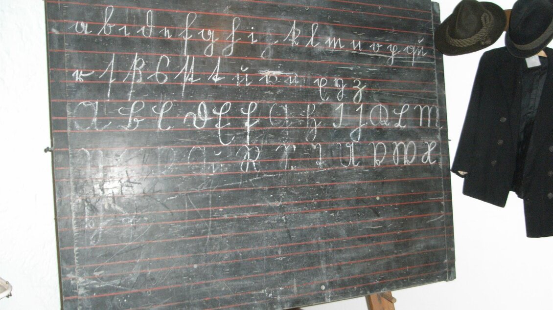 Eine alte Tafel mit handgeschriebenen Buchstaben in Kursivschrift. Im Hintergrund hängen einige Hüte und Kleidung. | © Stadtgemeinde Feldbach