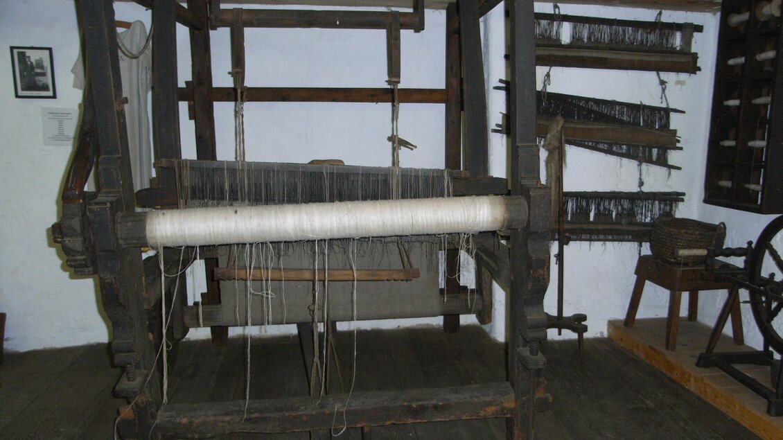 Ein alter Webstuhl aus Holz steht in einem Raum. Im Hintergrund sind weitere Webutensilien und Werkzeuge zu sehen. | © Stadtgemeinde Feldbach
