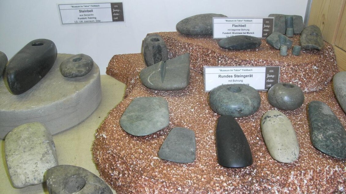 Eine Sammlung von Steinen und Werkzeugen aus der Steinzeit, präsentiert auf einem dekorativen Untergrund. Die Objekte sind unterschiedlich geformt und aus verschiedenen Materialien gefertigt. | © Stadtgemeinde Feldbach