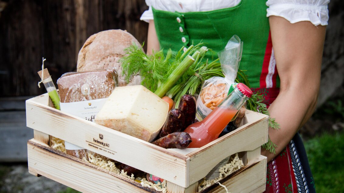 Ein Korb mit frischen Lebensmitteln, darunter Brot, Käse, Wurst und Gemüse. Die Person trägt ein traditionelles bayerisches Outfit. | © Heimatgold