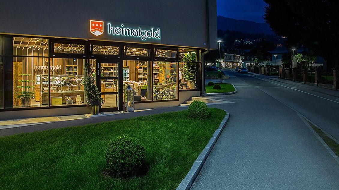 Ein Geschäft mit dem Namen "heimatgold" ist in der Dämmerung beleuchtet. Die Umgebung zeigt eine ruhige Straße und gepflegte grüne Rasenflächen. | © Heimatgold
