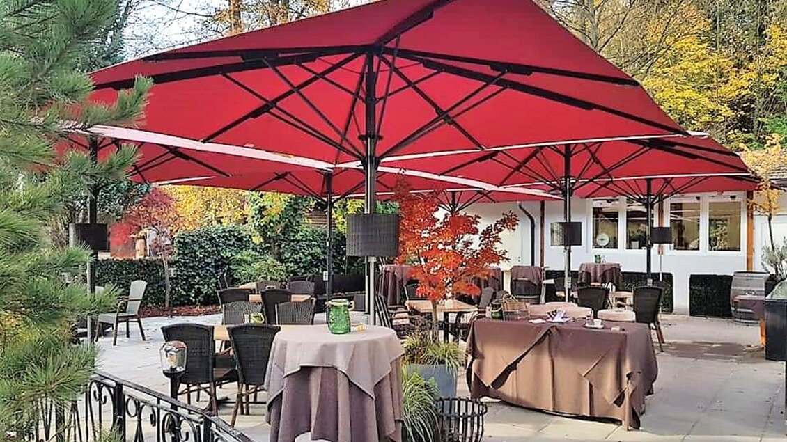 Ein gemütlicher Außenbereich eines Restaurants mit roten Sonnenschirmen. Die Tische sind elegant gedeckt und von Bäumen umgeben. | © Heiningers Schlemmerei