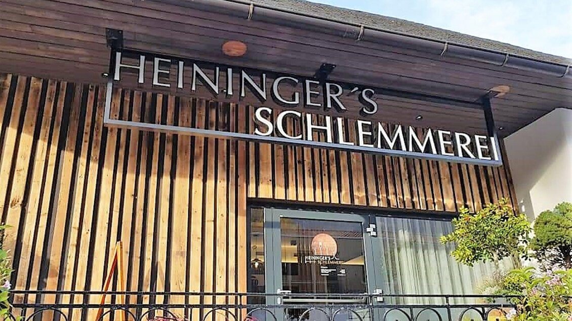 Ein einladendes Restaurant mit dem Namen "Heininger's Schlemmerrei". Die Fassade ist aus Holz und hat große Fenster, die einen Blick ins Innere ermöglichen. | © Heiningers Schlemmerei 
