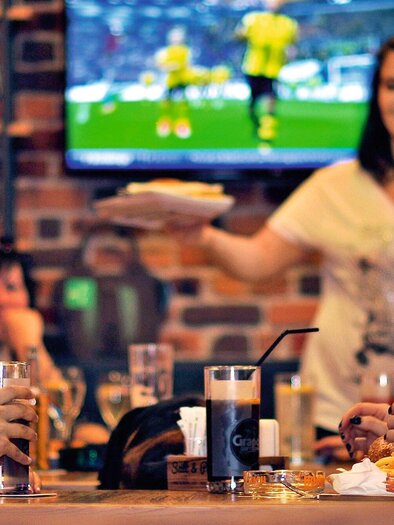 A sociable group of people sits in a restaurant enjoying drinks and food. In the background, a football game is playing on the television. | © HEINZ