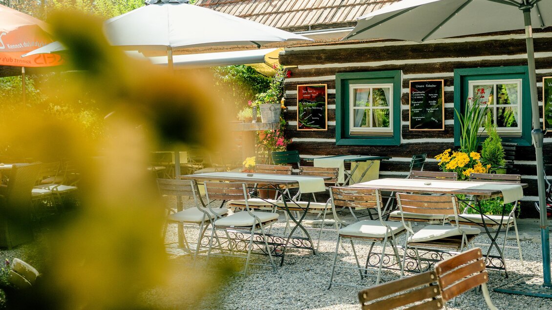 Ein gemütlicher Außenbereich mit Tischen und Stühlen unter Sonnenschirmen. Im Hintergrund steht ein rustikales Holzgebäude, umgeben von Blumen und Pflanzen. | © Region Graz - Mias Photoart