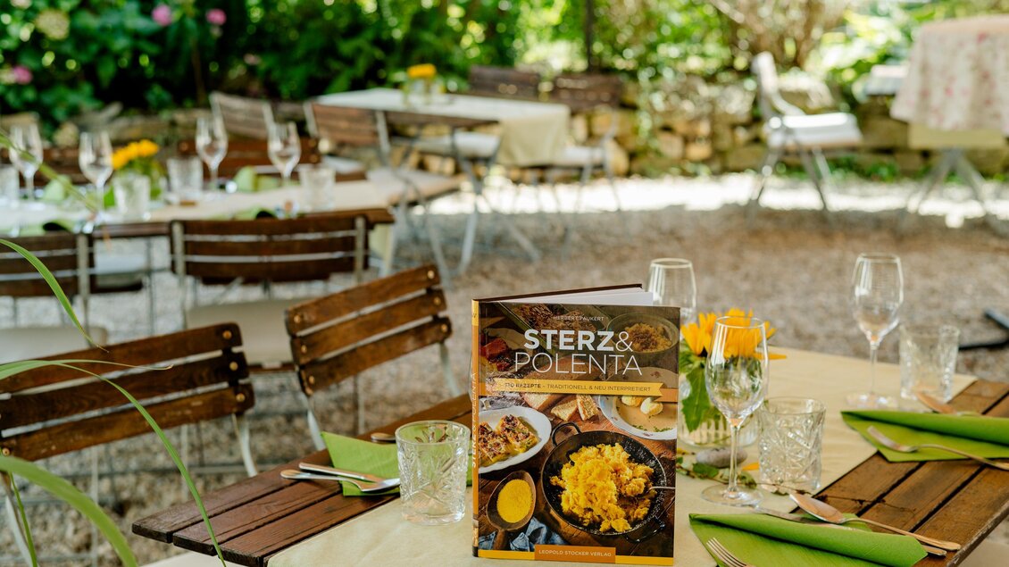 Ein einladender Garten mit Tischen und Stühlen, dekoriert mit Blumen. Im Vordergrund liegt ein Menü mit dem Titel „STERZ & POLENTA“. | © Region Graz - Mias Photoart