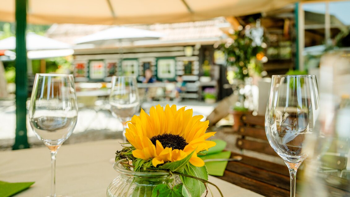 Ein gedeckter Tisch mit Weingläsern und einer Sonnenblume in einer Vase. Im Hintergrund sind weitere Tische und eine Menütafel sichtbar. | © Region Graz - Mias Photoart