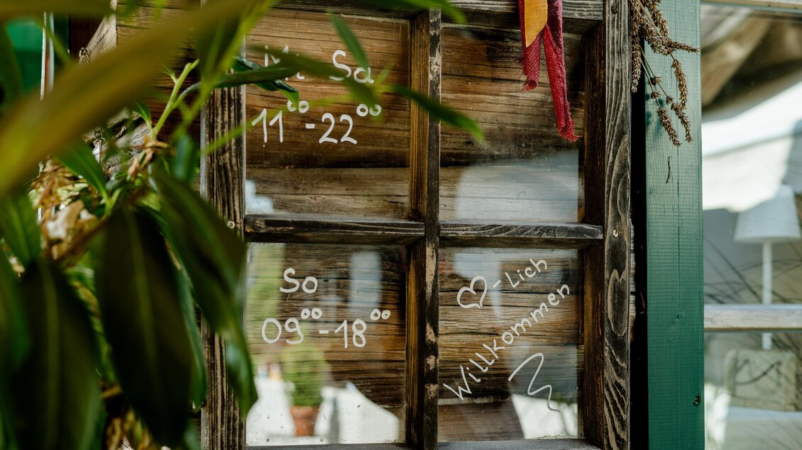 Ein Fenster mit einem Holzrahmen und Öffnungszeiten. Auf der Scheibe steht "Willkommen" und die Zeiten für Samstag und Sonntag. | © Region Graz - Mias Photoart