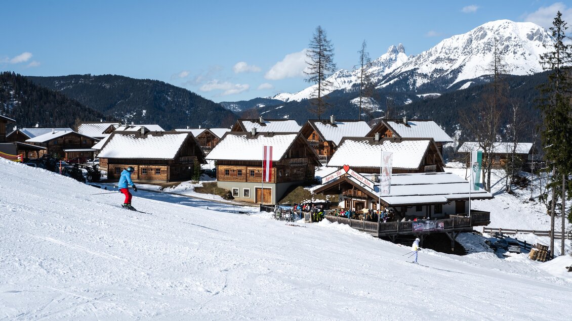Eine alpine Landschaft mit schneebedeckten Hütten und einem Skifahrer, der die Piste hinunterfährt. Im Hintergrund sind hohe Berge und blauer Himmel zu sehen. | © Adam Stocker