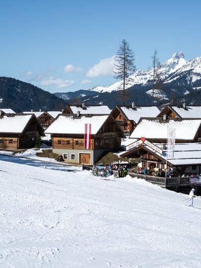 Eine alpine Landschaft mit schneebedeckten Hütten und einem Skifahrer, der die Piste hinunterfährt. Im Hintergrund sind hohe Berge und blauer Himmel zu sehen. | © Adam Stocker