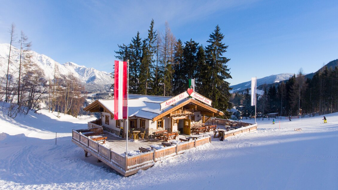 Eine gemütliche Hütte in den Bergen, umgeben von Schnee und Bäumen. Im Hintergrund erstrecken sich majestätische Berge unter einem klaren blauen Himmel. | © Strasser