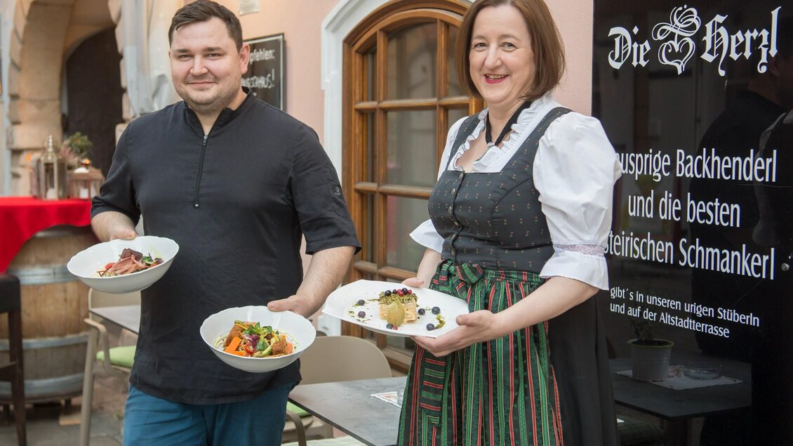 Ein Mann in schwarzem Hemd und eine Frau im Dirndl stehen vor einem Restaurant und halten Teller mit Speisen. Im Hintergrund ist ein Schild mit dem Namen "Die Herzl" zu sehen, das auf die Spezialitäten des Hauses hinweist. | © Herzl Weinstube
