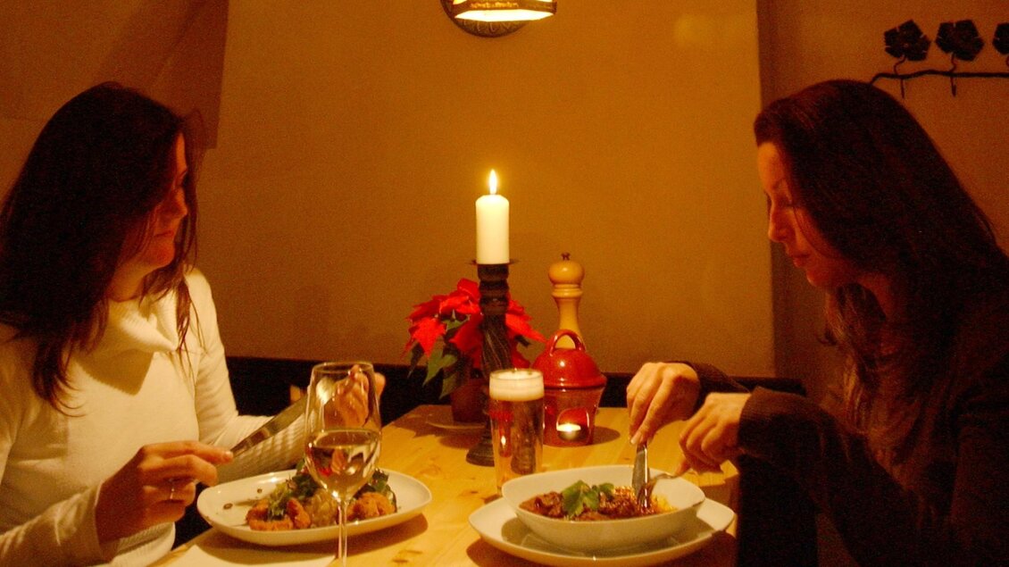 Zwei Personen genießen ein entspanntes Abendessen in einem rustikalen, gemütlich beleuchteten Restaurant. Auf dem Tisch stehen ein Glas Weißwein und ein Bier, während die Gäste in einer entspannten Atmosphäre ihr Gericht essen, umgeben von Kerzenlicht und Dekoration | © Herzl Weinstube