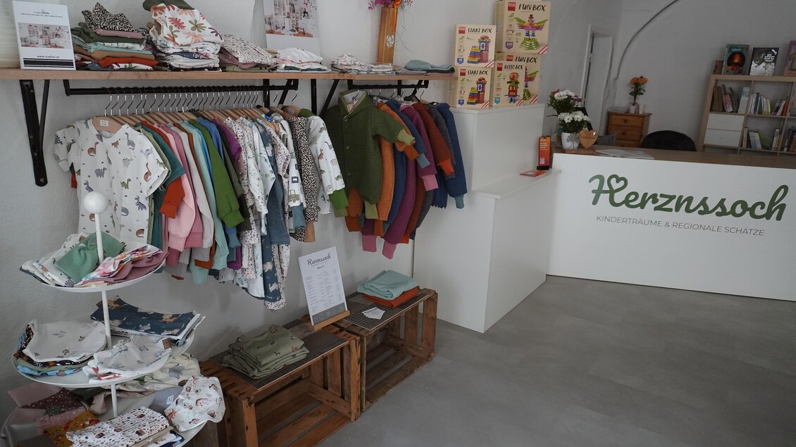 Ein gemütliches Geschäft mit bunten Kinderbekleidungen an einer Kleiderstange. Im Hintergrund ist der Empfangsbereich mit dem Logo „Herznssoch“ zu sehen. | © Denise Siedler
