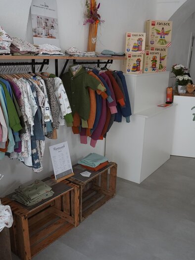 A cozy shop with colorful children's clothing on a clothing rack. In the background, the reception area with the logo "Herznssoch" can be seen. | © Denise Siedler